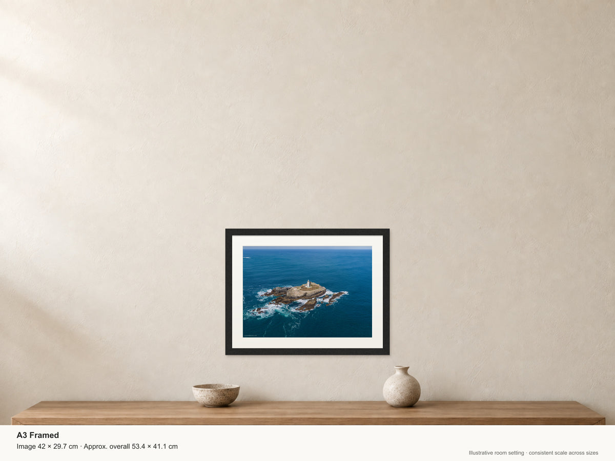 Close aerial view of Godrevy Lighthouse, enclosing stone walls and rugged island rocks surrounded by blue sea and white surf — A3 22mm black stained frame and 35mm mount. Illustrative room view with dimensions.
