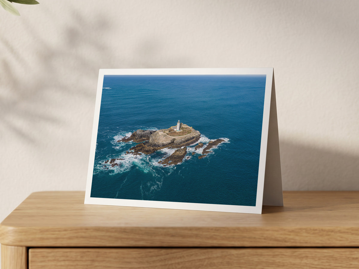 Close aerial view of Godrevy Lighthouse, enclosing stone walls and rugged island rocks surrounded by blue sea and white surf, shown on an A5 greeting card with a white border on an oak sideboard