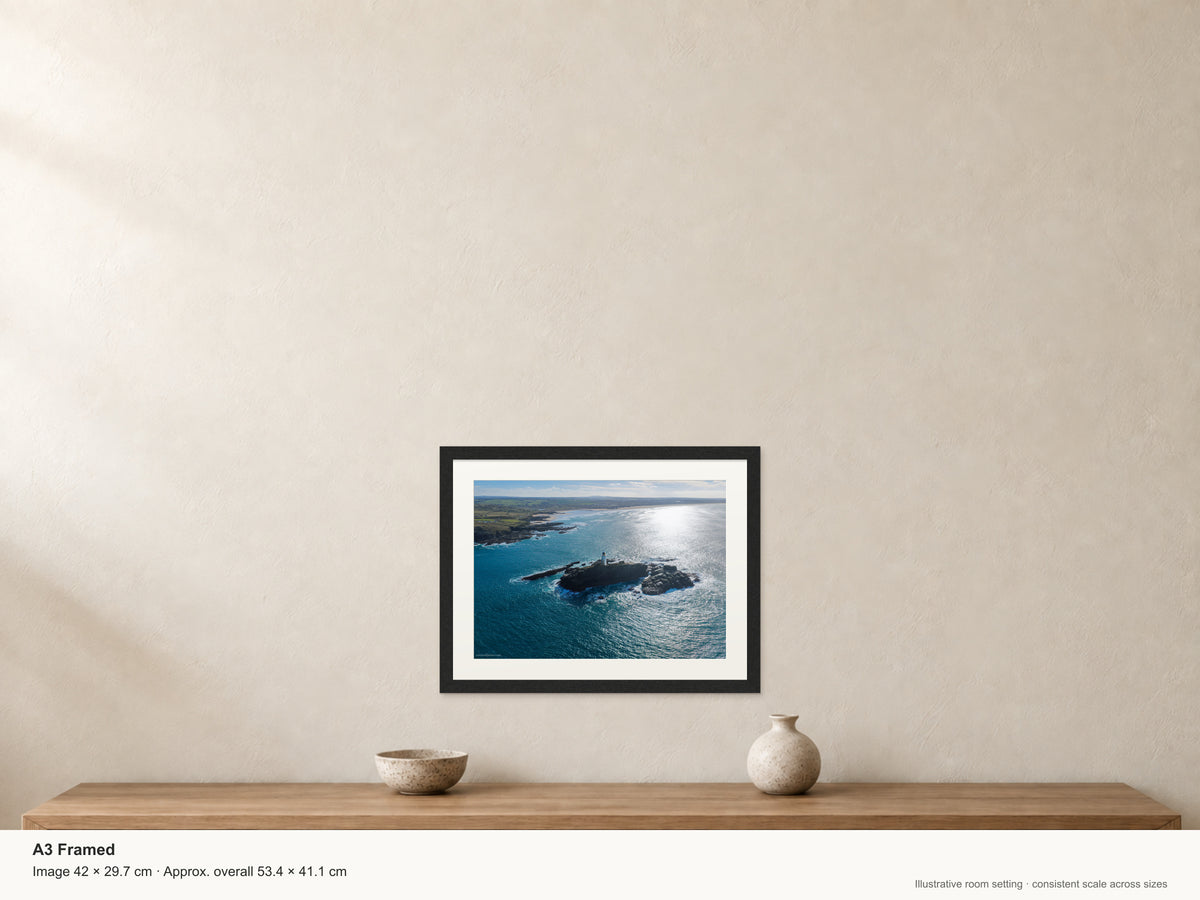 Godrevy Lighthouse on its rocky island with brilliant autumn sunlight on the sea and the coast towards Gwithian behind it — A3 22mm black stained frame and 35mm mount. Illustrative room view with dimensions.