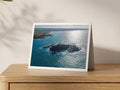 Godrevy Lighthouse on its rocky island with brilliant autumn sunlight on the sea and the coast towards Gwithian behind it, shown on an A5 greeting card with a white border on an oak sideboard