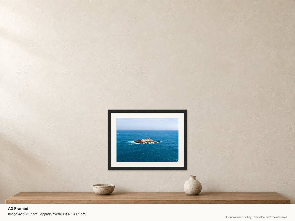 Godrevy Lighthouse isolated on its rocky island in a wide expanse of blue sea beneath a pale sky — A3 22mm black stained frame and 35mm mount. Illustrative room view with dimensions.