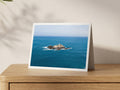 Godrevy Lighthouse isolated on its rocky island in a wide expanse of blue sea beneath a pale sky, shown on an A5 greeting card with a white border on an oak sideboard