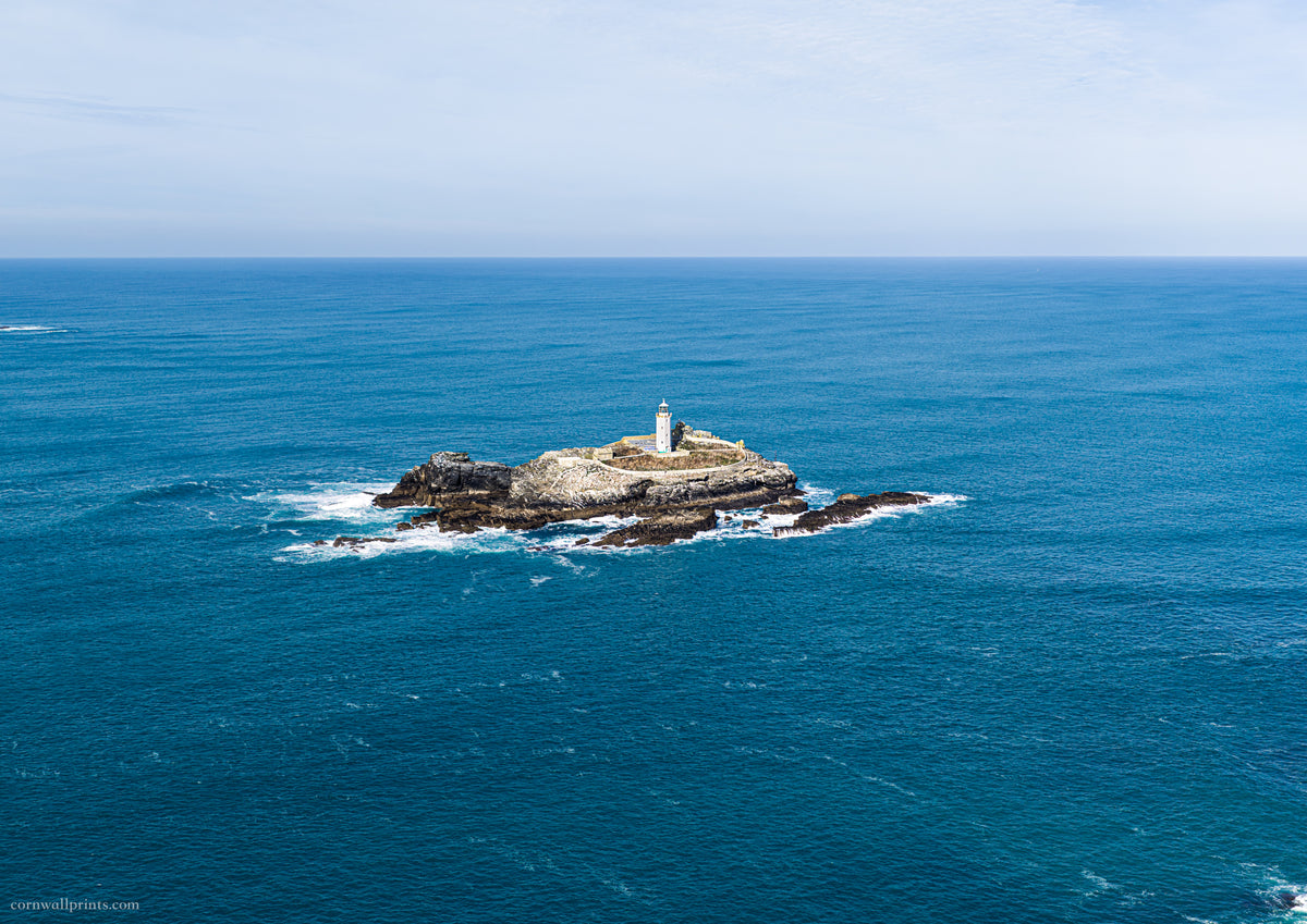 Godrevy Lighthouse isolated on its rocky island in a wide expanse of blue sea beneath a pale sky