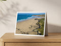 Expansive Gwithian beach with golden sand, textured cliffs, clean offshore surf and surfers, looking north towards Godrevy Lighthouse, shown on an A5 greeting card with a white border on an oak sideboard