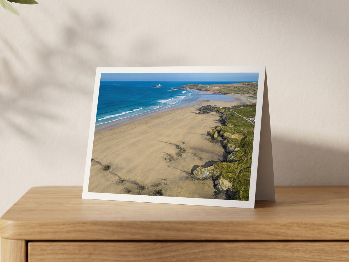 Expansive Gwithian beach with golden sand, textured cliffs, clean offshore surf and surfers, looking north towards Godrevy Lighthouse, shown on an A5 greeting card with a white border on an oak sideboard