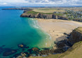 Aerial view from offshore looking north across Mawgan Porth at low tide, with turquoise-green water, golden sand and rugged cliffs