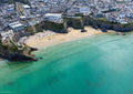 Aerial view of Great Western Beach, Newquay, with clear green-blue water, swimmers, surfers and the town above the cliffs at mid tide