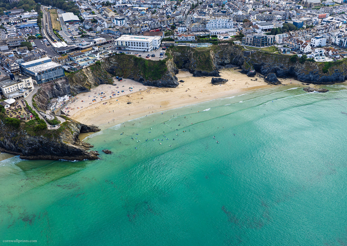 Aerial view of Great Western Beach, Newquay, with clear green-blue water, swimmers, surfers and the town above the cliffs at mid tide