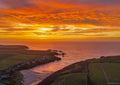 Brilliant orange and gold sunset over Porthcothan Bay, with light reflected across the sea and wet sand beneath the cliffs.