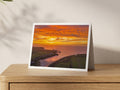 Porthcothan Inferno in the Sky greeting card: orange and gold sunset over the bay, printed with a white border on an A5 card standing on an oak sideboard.