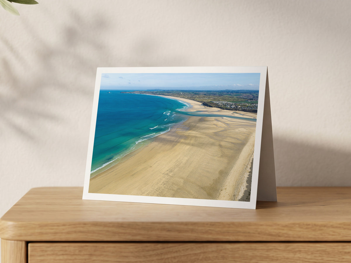 Porthkidney Sands at mid-to-low tide, looking across Hayle Estuary mouth along the expansive beaches to distant Godrevy Lighthouse, shown on an A5 greeting card with a white border on an oak sideboard