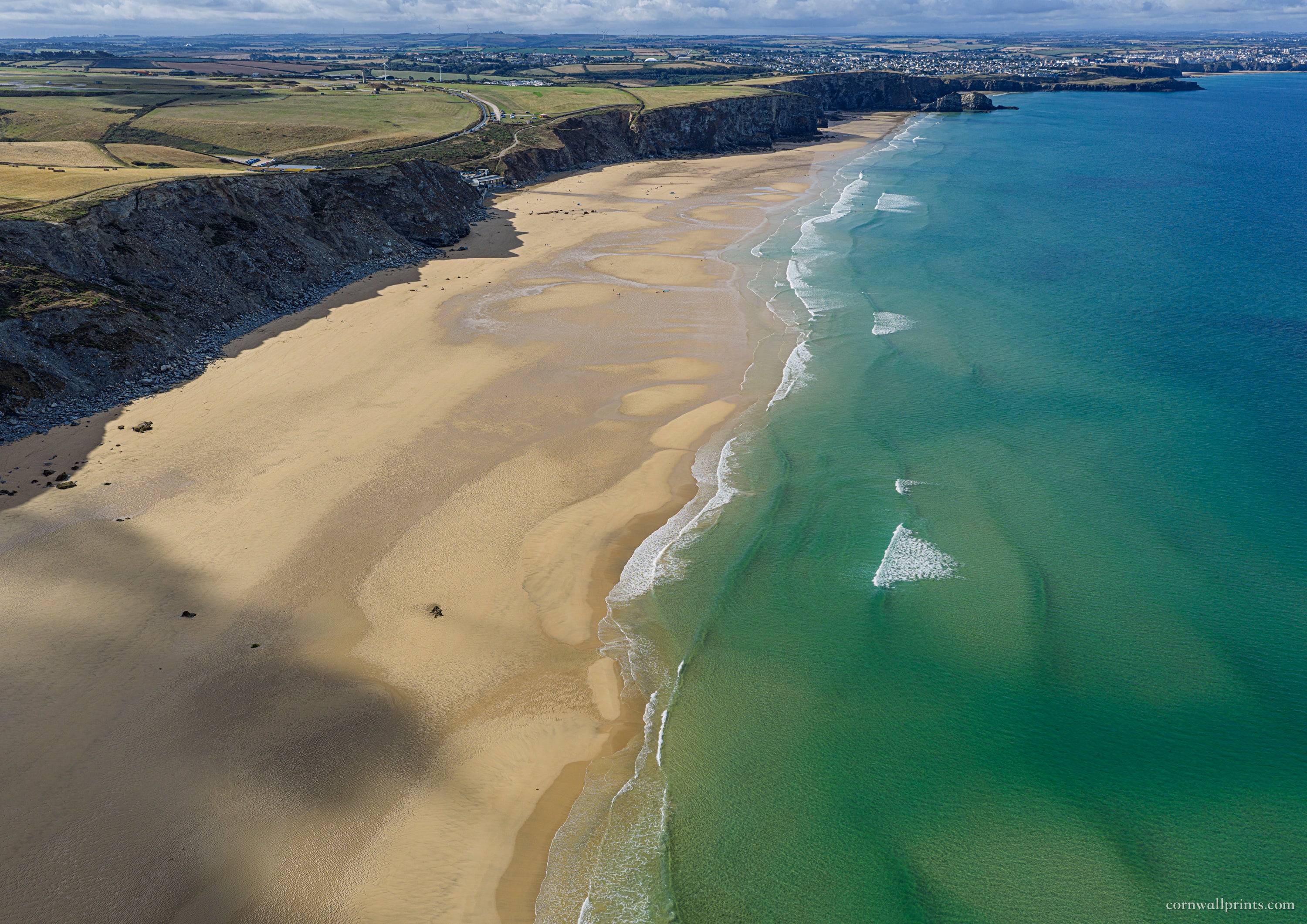 Aerial view looking south along Watergate Bay with turquoise water, low-tide sand, rugged cliffs and Newquay in the distance