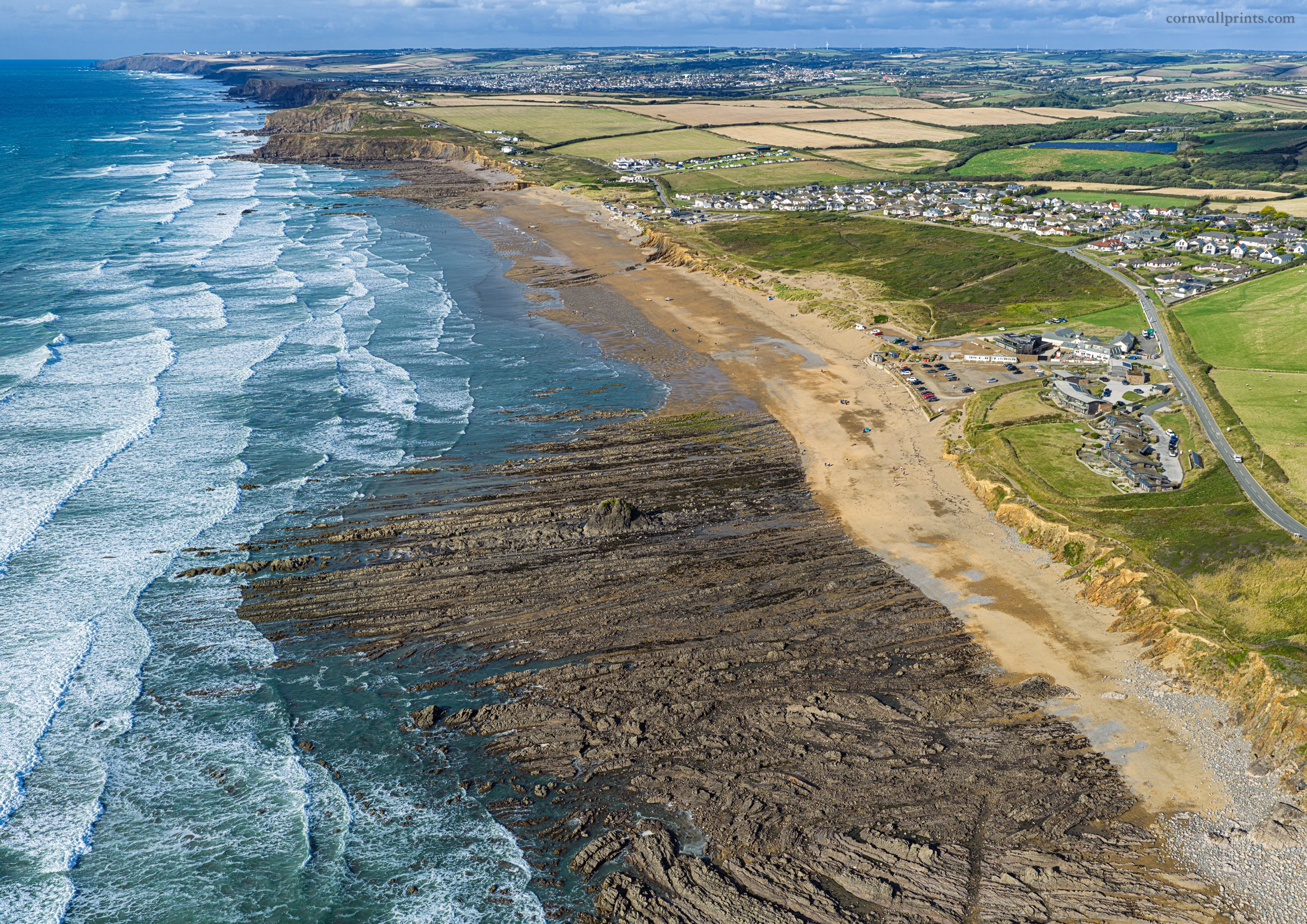 Aerial view from the south end of Widemouth Bay looking north towards Bude, with wind-driven surf, exposed reefs and the beachside village