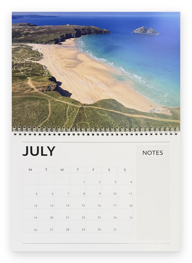 Printed Cornwall calendar 2027 open at July, a wide sandy beach and headland above the month grid