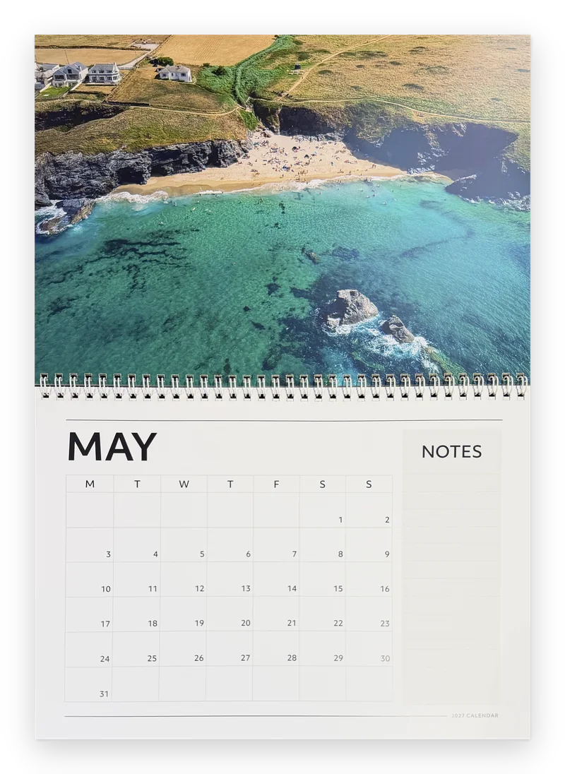 Printed Cornwall calendar 2027 open at May, a turquoise cove below clifftop houses above the month grid